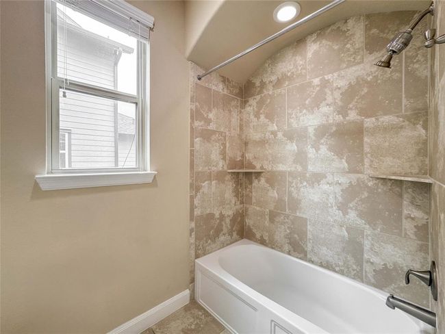 This bathroom features a modern tub and shower combo with elegant beige tile surround, a built-in shelf, and a recessed light above. A window provides natural light, enhancing the airy feel. | Image 43