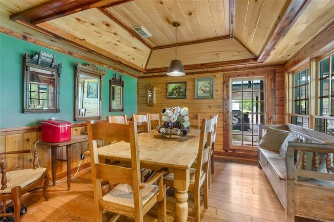 This cozy dining area features warm wood paneling, a rustic wooden table with matching chairs, and large windows that let in natural light. The room is accented with mirrors, artwork, and a pendant light, creating a welcoming and homey atmosphere. | Image 19
