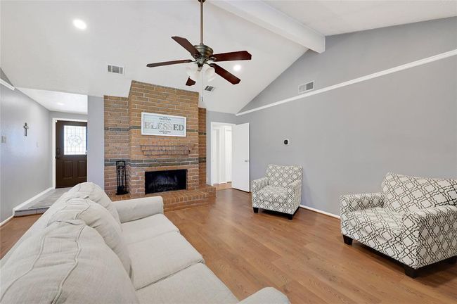 This inviting living room features a high vaulted ceiling with a ceiling fan, a cozy brick fireplace. The space is bright and open, with neutral gray walls perfect for relaxation and entertaining. | Image 6