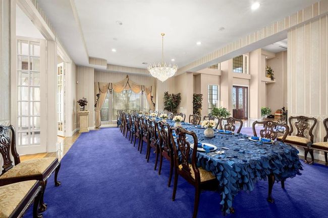 This is a spacious, formal dining room featuring a long table with elegant settings, a luxurious chandelier, high ceilings, and ample natural light from large windows. The room is accented by a striking purple carpet and has a grand staircase leading to the second level, suggesting a sophisticated space ideal for entertaining. | Image 7