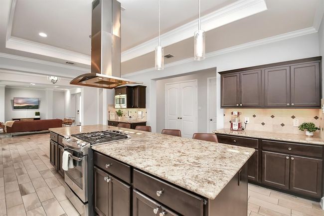 This spacious kitchen features a large island with granite countertops, modern appliances, and ample dark wood cabinetry. It flows seamlessly into an open living area, perfect for entertaining. | Image 8