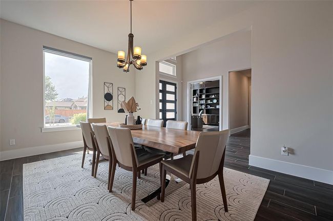 Bright dining area with modern chandelier, large window, and stylish decor. Features a wooden table with six chairs on a patterned rug. Open layout leads to a hallway and a glimpse of a cozy office space. | Image 7