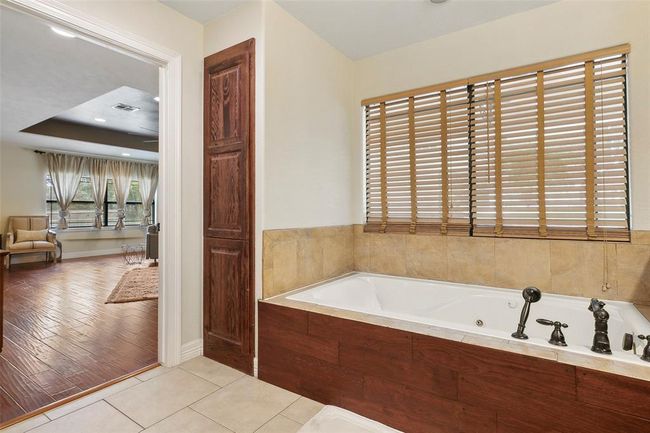 This photo showcases a cozy bathroom with a large corner bathtub, tiled walls, and wooden blinds. It opens into a spacious living area with wood flooring and large windows, offering plenty of natural light. | Image 30