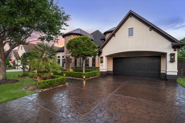 The residence boasts a blend of stone and stucco finishes, accented by striking rooflines and dark architectural shingles for a timeless European-inspired aesthetic. The extra-wide stamped concrete driveway offers ample parking and leads to a spacious 3-car tandem garage with a rich, dark wood carriage-style door. Inside the garage, premium features await, including finished floors, insulated walls, a convenient side door to the backyard, and an automatic garage door opener. | Image 4