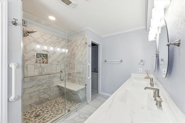 This primary bathroom features a spacious, modern design with a large glass-enclosed shower showcasing elegant marble tiles and a built-in bench. The double vanity with sleek faucets and ample counter space adds functionality and style. | Image 26