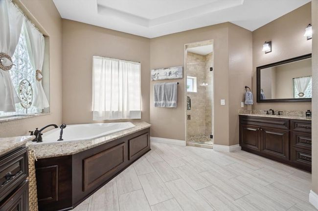 This spacious bathroom features a luxurious soaking tub, a separate glass-enclosed shower, and elegant dark wood cabinetry. The space is brightened by natural light from the windows, complemented by modern fixtures and neutral tones for a relaxing ambiance. | Image 10