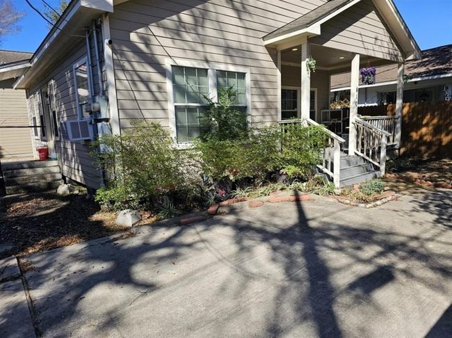 This photo shows the front of a cozy, single-story home with beige siding. It features a small porch with steps, surrounded by landscaped greenery and a concrete driveway. The porch has a railing and hanging plants, adding charm to the entrance. | Image 4