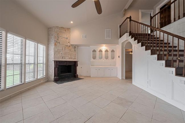 This spacious living room features a dramatic stone fireplace with ornate wood mantel, high ceilings, and built-in cabinetry for added storage and display. A curved staircase with dark wood railings adds architectural interest and a grand focal point. | Image 11
