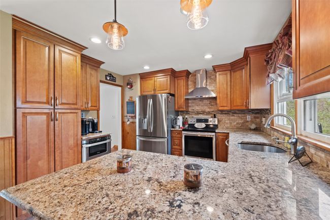 Kitchen featuring light stone countertops, brown cabinetry, stainless steel appliances, a sink, and wall chimney exhaust hood | Image 9