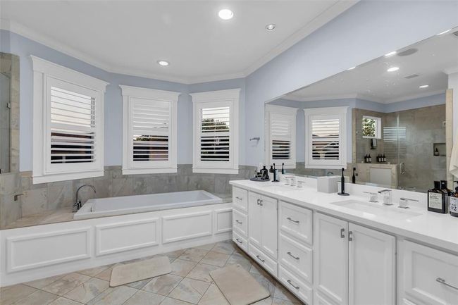 This Spa-like Primary bathroom features a spacious layout with a large jetted tub, dual sinks, a separate shower and ample counter space. It has natural light from multiple windows with shutters, complemented by a neutral tile floor and elegant cabinetry. | Image 21