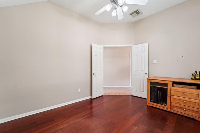 This room features warm hardwood flooring, neutral walls, and a ceiling fan with lighting. It includes double doors leading to another area and a wooden cabinet against one wall, offering a versatile space for various uses. | Image 12