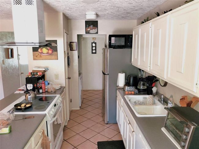 This kitchen features a galley layout with light-colored cabinetry and countertops. It includes a modern stovetop, oven, and a stainless steel refrigerator. The tiled floor and ample counter space provide functionality, while decorative touches add warmth. | Image 7