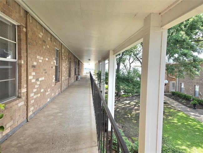 This photo shows a long, covered outdoor corridor with brick walls and a railing overlooking a landscaped area with trees and grass. It's part of a residential building complex, providing a view of a neighboring building and a pathway below. | Image 20