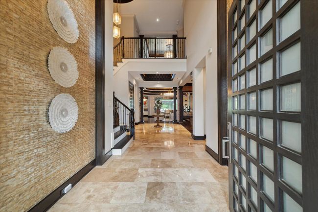 Welcome to a showstopping entrance where a soaring two-story foyer is wrapped in shimmering mother of pearl tiles, casting a soft iridescence throughout the space. The dramatic accent walls adorned with artistic décor sets the tone for refined luxury living. Gleaming travertine floors extend through the entry, while the soaring ceiling showcases a sampling of the $250, 000 lighting upgrades found throughout the home, which blends grandeur with warmth in this stunning entryway. | Image 5