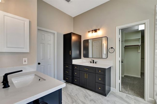 This modern bathroom features dual sinks with sleek black fixtures, elegant cabinetry, and a spacious walk-in closet. The neutral tones and marble-style flooring add a touch of luxury. | Image 34