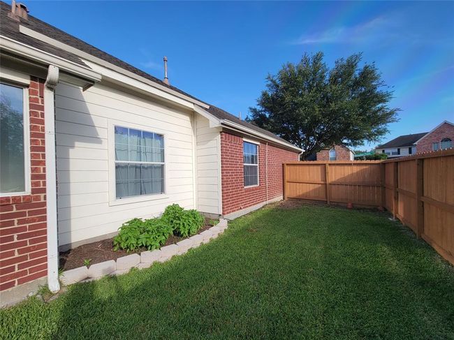 This photo shows a cozy backyard with a well-maintained lawn, bordered by a wooden fence. The exterior features a combination of red brick and white siding, with a small garden bed under the window. It's a private, inviting outdoor space. | Image 32