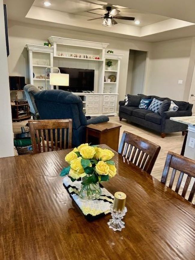 This cozy living area features a dining table with a floral centerpiece, a comfortable seating arrangement with a sofa and armchair, and room for an entertainment unit. The room has a warm ambiance with wood-look tile flooring and recessed lighting. | Image 11