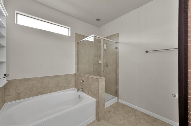 This bathroom features a modern design with a spacious bathtub and a separate glass-enclosed shower. It has neutral tiles, a horizontal window for natural light, and a towel bar. | Image 20