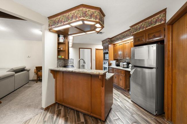 Kitchen featuring stainless steel appliances, brown cabinetry, a peninsula, light wood-type flooring, and light stone countertops | Image 13