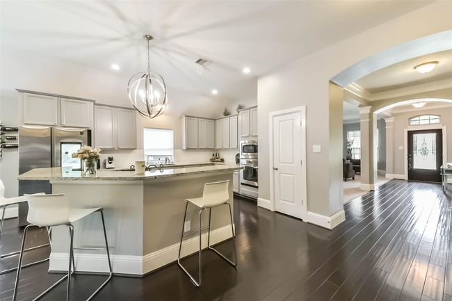 Showcasing a modern kitchen with sleek dark hardwood floors, a spacious island with bar seating, stainless steel appliances, and white cabinetry. The open layout flows into a bright entryway with elegant archways. | Image 14