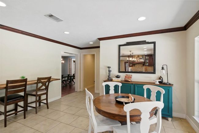This cozy dining area features a round wooden table with white chairs, a teal sideboard with decorative items, and a large mirror. The space is bright with recessed lighting and has tiled flooring, offering a welcoming atmosphere. | Image 18