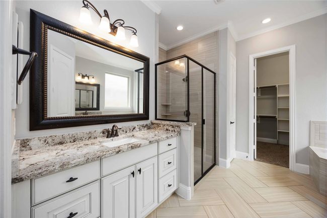 This bathroom features a modern design with a granite-topped vanity, elegant light fixtures, and a large framed mirror. It includes a glass-enclosed shower with stylish tile work and an adjacent walk-in closet. | Image 22