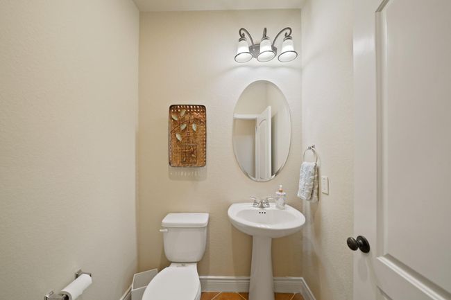 This is a compact, well-lit bathroom with a modern design. It features a pedestal sink, oval mirror, toilet, and stylish light fixture. The neutral color scheme and decorative wall art add a cozy touch. | Image 24