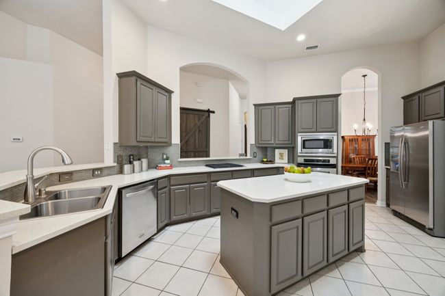 Natural light pours in from a central skylight, highlighting the soft gray cabinetry, wraparound breakfast bar, and easy access to both formal and casual dining areas. | Image 6