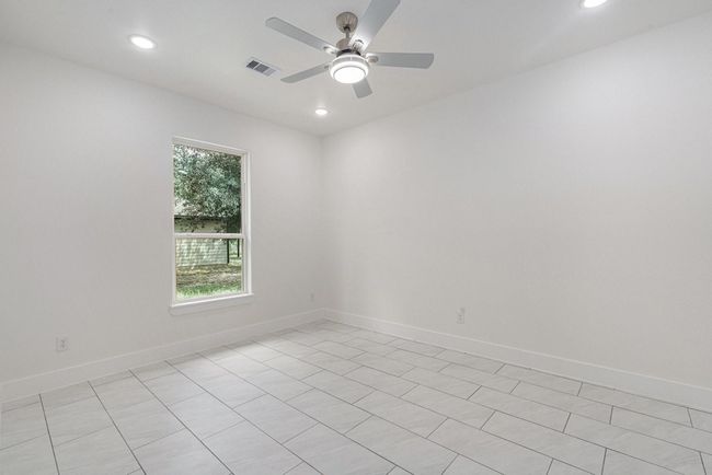 Bright, spacious room with white walls and tiled flooring. It features a ceiling fan and recessed lighting, with a window providing natural light and a view of greenery outside. | Image 34