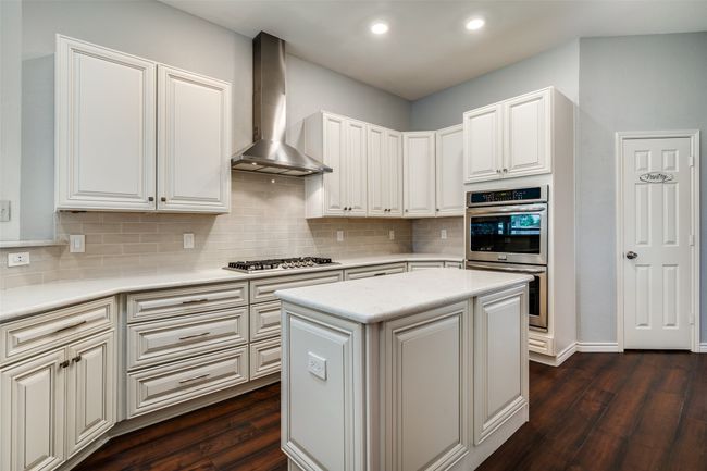 Kitchen with dark wood-style flooring, decorative backsplash, wall chimney exhaust hood, a kitchen island, and white cabinets | Image 13