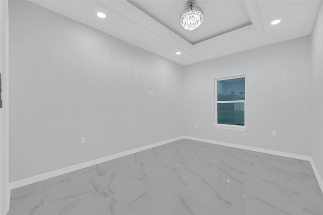 Bright room with modern design, featuring light gray walls and glossy marble-like tile flooring. A large window allows natural light, while recessed lighting and a stylish chandelier add elegance. The tray ceiling adds a touch of sophistication. | Image 27