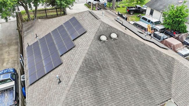 This is an aerial view of a property featuring a shingled rooftop with solar panels installed, suggesting energy efficiency and potential savings on utility bills. Surrounding the property are vehicles and another building, indicating a suburban setting. | Image 12