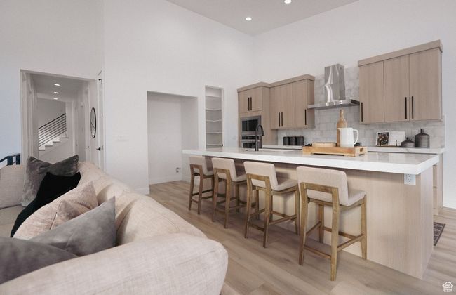 Kitchen with wall chimney range hood, a high ceiling, light wood-style floors, light countertops, and a kitchen breakfast bar | Image 5