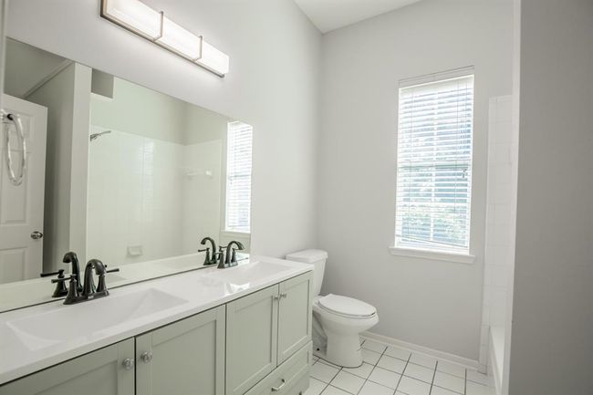 Bright and modern bathroom featuring a double vanity with sleek fixtures, a large mirror, and natural light from a window. The space includes a bathtub and white tile flooring, creating a fresh and clean atmosphere. | Image 36