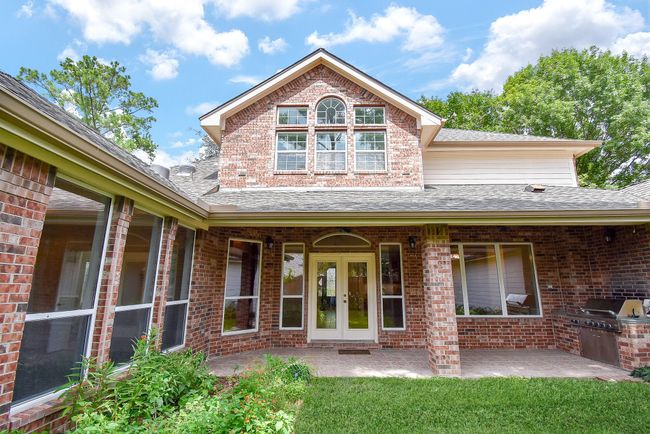 This photo showcases the back of a charming brick home with a spacious covered patio, large windows, and a built-in outdoor grill. The lush green lawn and surrounding trees add a serene touch. | Image 44