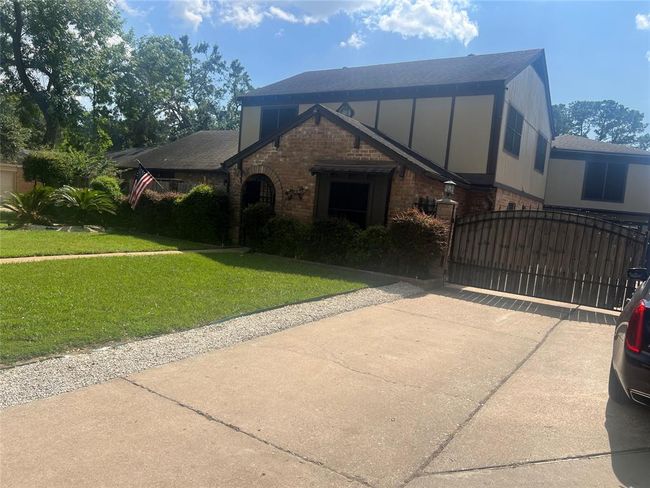 This photo shows a two-story brick house with a well-maintained lawn and a driveway leading to a gated entrance. The home features a combination of brick and siding, with mature trees and manicured shrubs in the front yard, creating a welcoming and private atmosphere. | Image 5