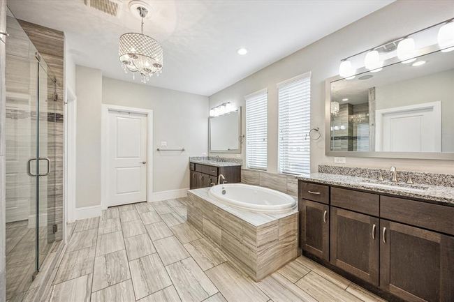 This bathroom features a modern design with a spacious layout, including a luxurious bathtub, dual sinks with granite countertops, dark wood cabinetry, and a glass-enclosed shower. The room is well-lit with large windows and an elegant chandelier, complemented by light tile flooring. | Image 20
