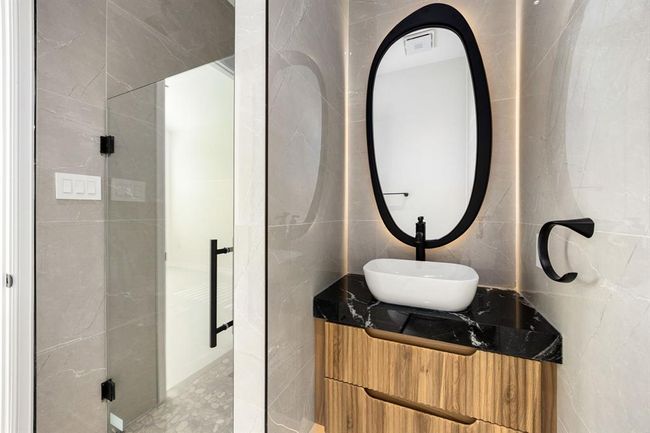 This stylish en suite bath features contemporary elegance with sleek gray tile walls, a bold black-framed mirror, and a striking vessel sink set atop a rich wood vanity with black marble countertop. A frameless glass shower and modern matte black fixtures complete the sophisticated, spa-like design. | Image 28