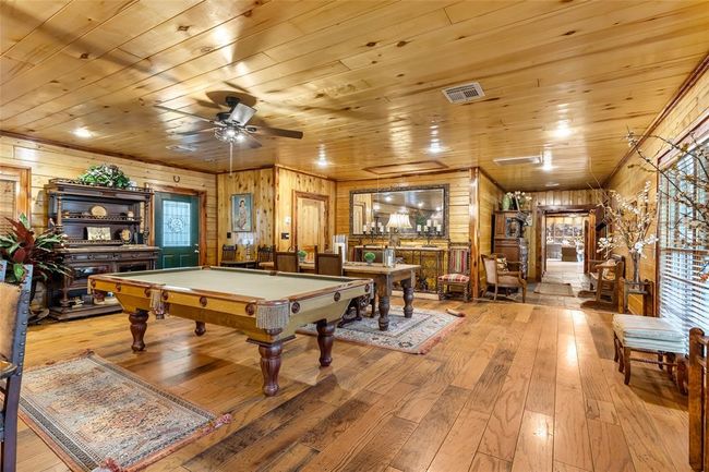 This photo showcases a spacious, rustic game room with wood-paneled walls and ceiling, featuring a billiards table at the center. The room has hardwood flooring, a ceiling fan, and is furnished with classic wooden furniture and decorative rugs, creating a cozy, inviting atmosphere. | Image 36