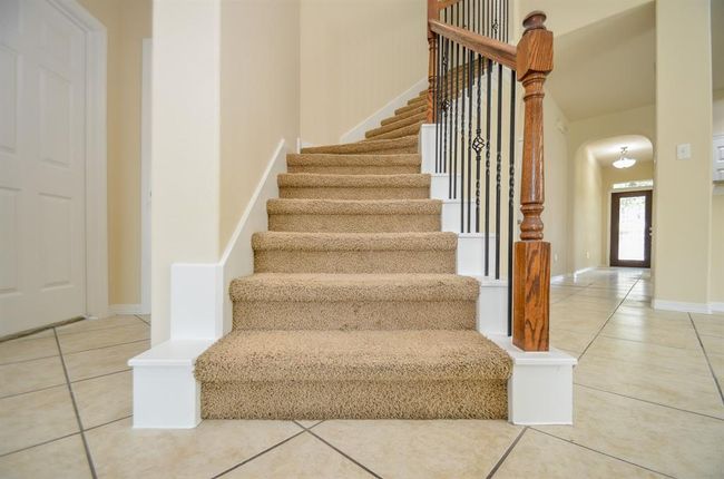 An inviting entryway and a carpeted staircase with elegant wooden railing details. The space is bright with neutral walls and tiled flooring, leading to a well-lit hallway. | Image 9