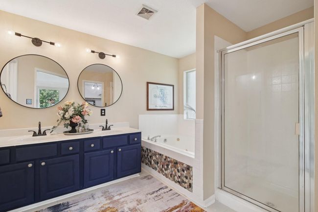 This bathroom features a double vanity with elegant dark blue cabinets, modern round mirrors, and stylish lighting. It includes a spacious glass-enclosed shower and a corner bathtub with decorative tile accents. A window provides natural light, enhancing the warm and inviting atmosphere. | Image 21