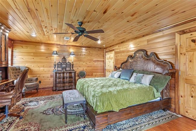 This cozy bedroom features rustic wood paneling on the walls and ceiling, a ceiling fan, and elegant wooden furniture. The room includes a plush bed with decorative pillows, a comfortable desk chair, and a patterned rug, creating a warm, inviting atmosphere ideal for relaxation. | Image 47