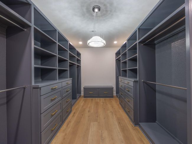 Indulge in a boutique-style walk-in closet with extensive built-in shelving, drawer systems, and dual hanging space—all wrapped in elegant slate blue tones and finished with gold hardware and a sparkling chandelier. | Image 6