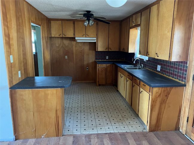 Kitchen with wooden walls, ceiling fan, dark countertops, light floors, and a sink | Image 8