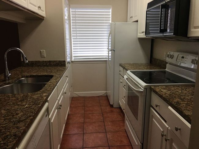 Kitchen with white appliances, white cabinets, dark tile patterned flooring, dark stone countertops, and a sink | Image 9