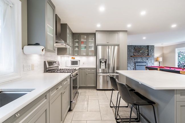 Kitchen featuring wall chimney exhaust hood, a kitchen bar, a stone fireplace, gray cabinetry, and stainless steel appliances | Image 10