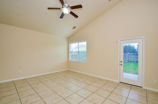Bright living room with high ceilings, neutral walls, and tile flooring. Features a ceiling fan and large window for natural light, plus a door leading to the backyard. | Image 16