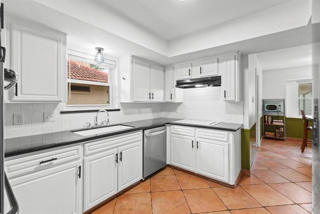 This kitchen features sleek white cabinetry, a modern stainless steel dishwasher, and granite countertops. The space is brightened by a window above the double sink and has terracotta floor tiles for a warm, inviting feel. An adjacent breakfast area is visible, enhancing the open layout. | Image 8
