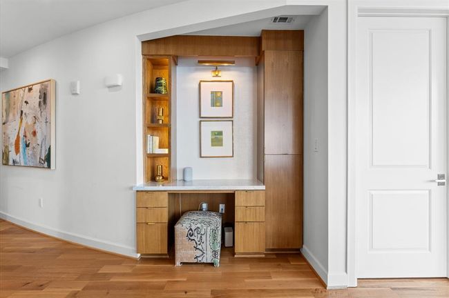 This photo showcases a modern built-in nook with wooden cabinetry, open shelving, and a marble surface, perfect for a small office or decorative space. It features contemporary artwork and a clean, minimalist design, accented by a plush ottoman and warm wood flooring. | Image 7