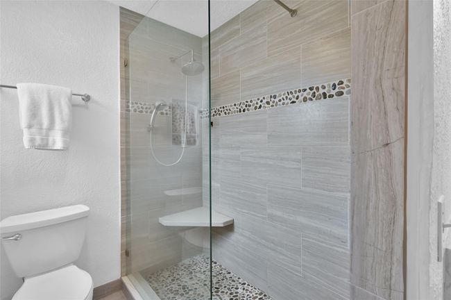 The Primary Bath continues with a frameless shower with rain showerhead and hand-held shower wand with temperature control located near the opening. Wood-look tile with pebble accents, product niche and sitting seat make showering a relaxing experience. | Image 18
